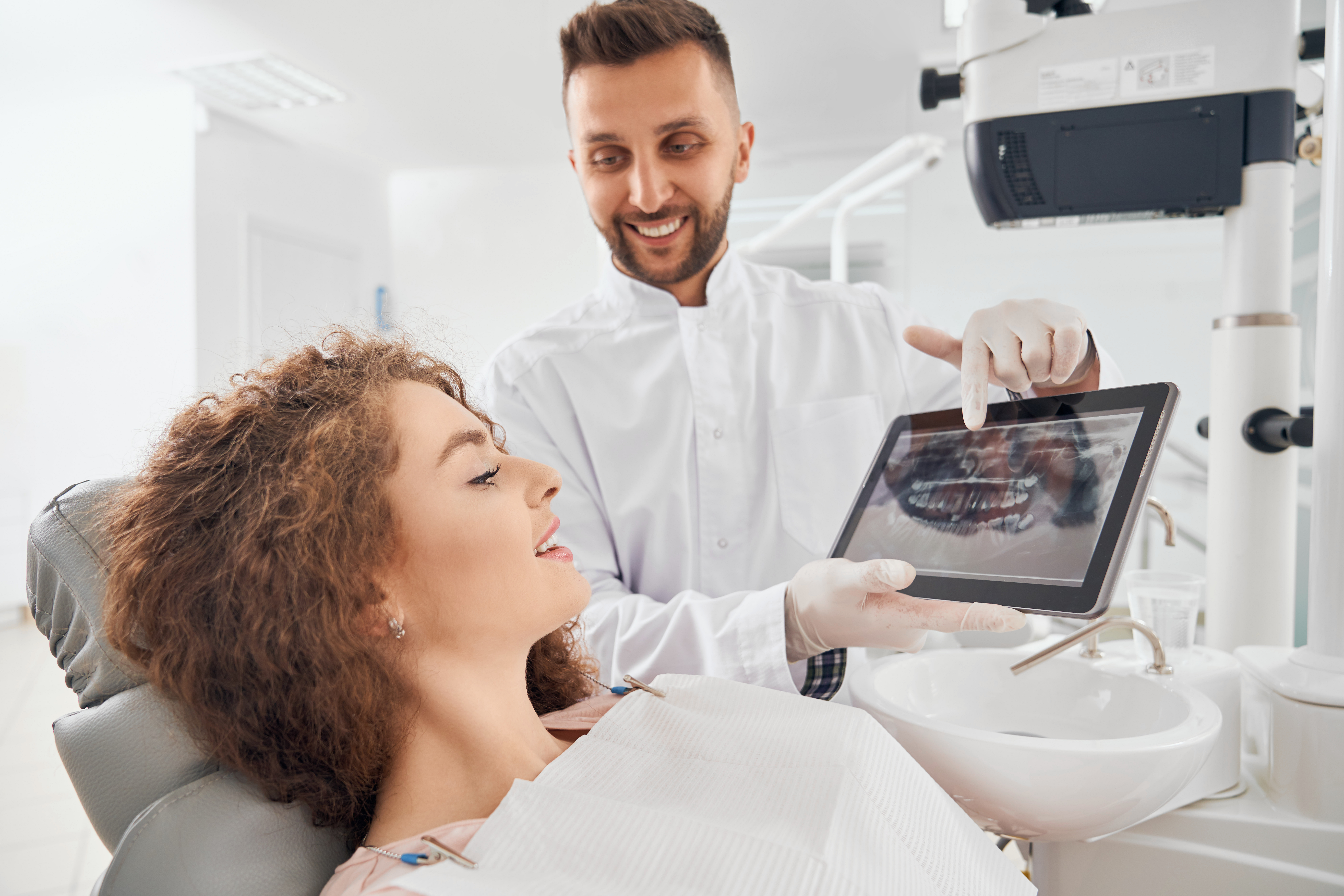 Handsome male doctor in white medical uniform and rubber gloves holding electronic tablet and showing beautiful female client x-ray pictures of her teeth. Concept of dentistry.