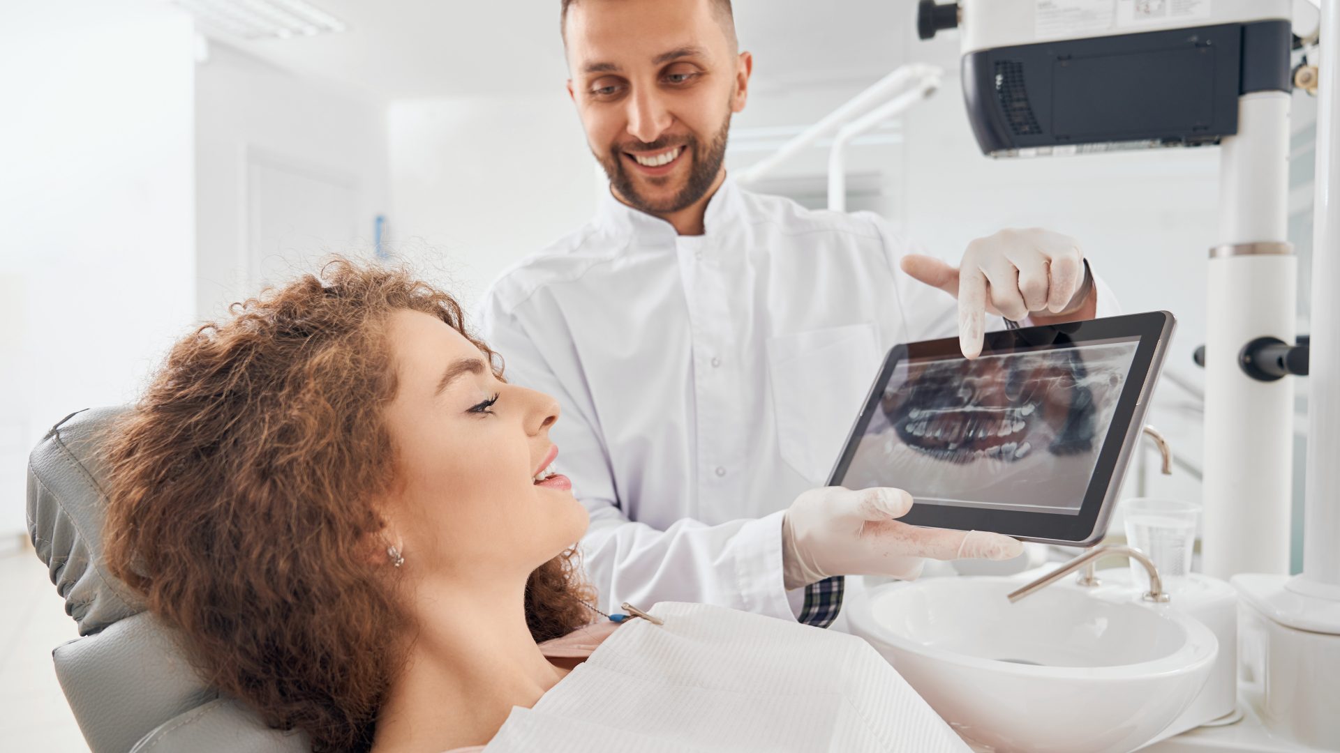 Handsome male doctor in white medical uniform and rubber gloves holding electronic tablet and showing beautiful female client x-ray pictures of her teeth. Concept of dentistry.